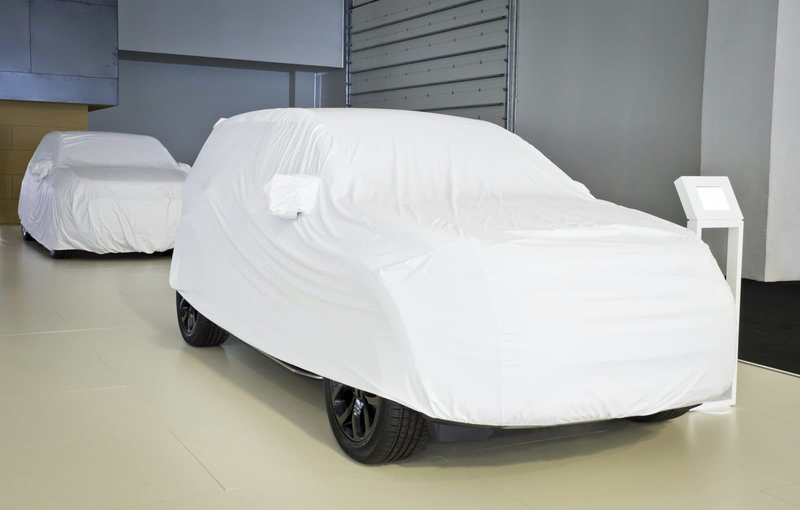 Two cars under white fabric covers in a bright indoor showroom