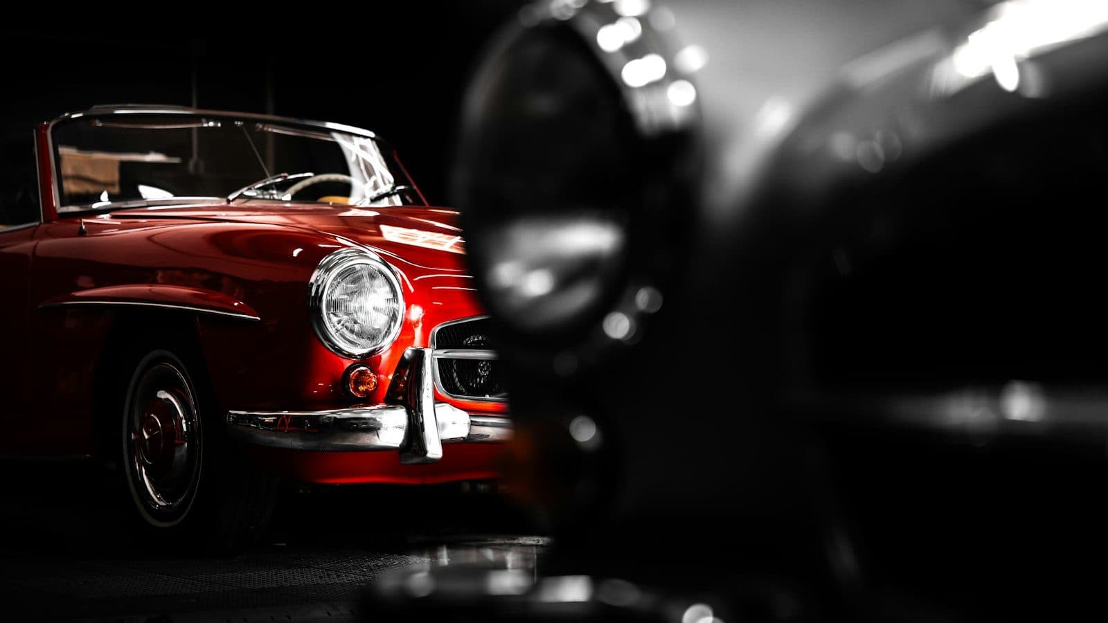 A red classic roadster parked in a dark garage, seen past a blurred chrome headlamp