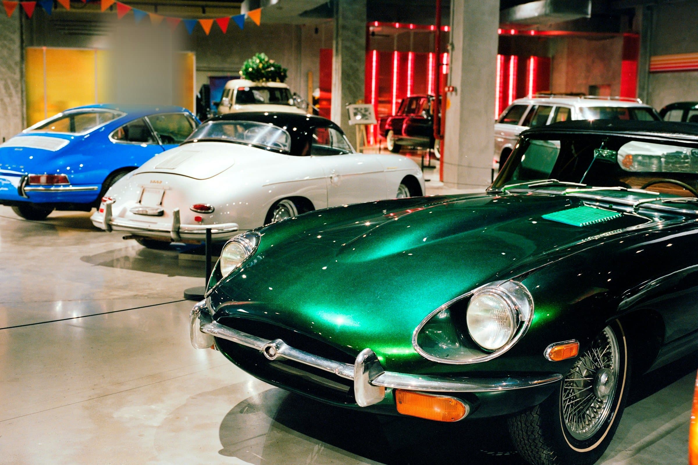 A green Jaguar E-Type in front of a blue Porsche 911 and a white Porsche 356, parked indoors